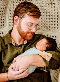 Dad holding newborn baby in boho high back chair for studio lifestyle newborn session
