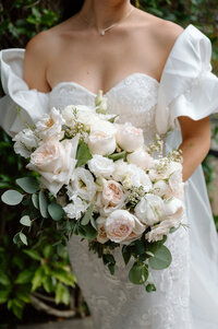 A close up of a brides bouquet as she holds it in front of her. The flowers are white roses, peonies and green leafs. 