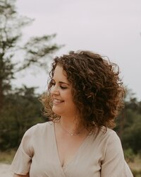 Woman with short curly brown hair smiling while looking to the side, wearing a beige V-neck dress outdoors with pine trees softly blurred in the background.