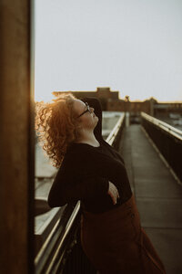 A rooftop senior session at golden hour . The sun is shining behind her as she leans against the railing, hand in her curly hair.