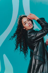 Curly-haired creative poses with hand raised in front of teal abstract mural – bold and artistic brand portrait by Mewphotovideo