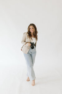 Branding photo for photographer, a blonde woman wearing jeans, white top, and olive green blazer while holding her phone and smiling at it