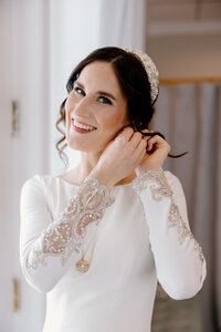 Catholic bride puts on her earrings and smiles at the camera as she's getting ready for her wedding Mass.