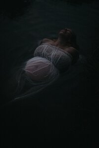 A pregnant woman is laying in water wearing a transparent white dress. Her pregnant belly is visible through the dress and she has her head back.