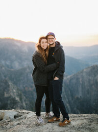 man and woman standing on cliff overlooking mountain valley