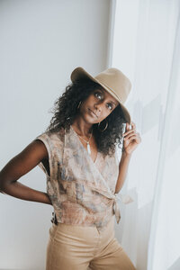 Fashionable woman with curly hair wearing a tan wide-brimmed hat, sleeveless patterned top, and tan pants, standing by a white curtain and gazing at the camera.