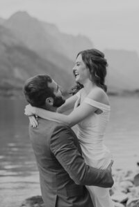 A bride laughs while her groom lifts her beside a serene mountain lake during their Glacier National Park elopement, captured in timeless black and white by Sydney Breann Photography.