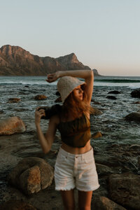 young woman wearing a bucket hat stares off into the distance  with the mountains and  the ocean behind her