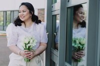 Bride standing outdoors holding a bouquet of white tulips, smiling as she looks at her reflection in a glass window.