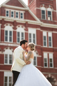 A bride and groom share a romantic kiss in front of a historic red brick building on the Auburn University campus. The groom, dressed in a white suit, embraces his bride, who wears a stunning ballgown with intricate lace details. This timeless wedding portrait captures the elegance and emotion of Alabama wedding photography.