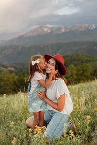 A woman in a red hat kneels in a grassy field dotted with yellow flowers, embracing a young girl in denim overalls who kisses her cheek. Against the backdrop of majestic mountains, this tender moment is beautifully captured by a Colorado Elopement Photographer.