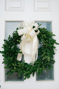 Greenery round wreath hanging on a white door with an off white ribbon 