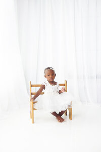 Baby girl wearing white dress sitting on tiny bench during mommy and me photoshoot in Franklin Tennessee photography studio