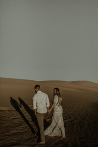 Couple walks hand in hand across the image with their shadows leading them across the sand.