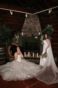 wo brides in elegant white wedding dresses posing in an editorial style near a rustic stone fireplace adorned with candles and floral decorations, set in a cozy cabin wedding venue—captured by a destination wedding photographer specializing in LGBTQ+ and Pacific Northwest weddings.