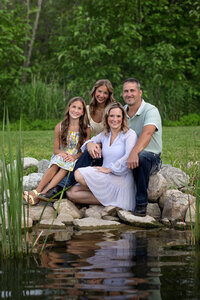 A family of four sits smiling on rocks on the edge of a pond