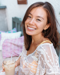 A woman is smiling while holding a drink with a straw. She is outside on a patio, wearing a white, lace-patterned top. Pillows are visible in the background, providing comfort as she enjoys her moment of relaxation as the copywriter of a Shreveport website design company.