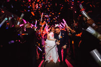 A couple exits their wedding through a colorful crowd of guests and light