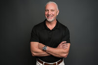  A business headshot of an older man with gray hair, smiling warmly while crossing his arms. He wears a black shirt against a dark gray background, exuding confidence, charisma, and a fun, approachable energy. Captured by Leslie Q Photography, this portrait showcases professionalism with personality.