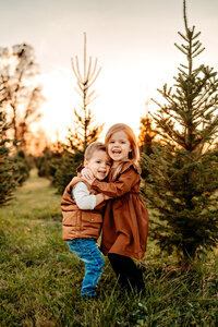 Brother and sister hugging each other at a christmas tree farm with sunset behind them