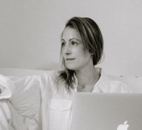 Black and white photo of a woman in a relaxed pose, wearing a loose white shirt and working on a MacBook, gazing off to the side in a calm, minimalist space.