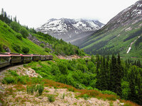 A scenic mountain railway in Alaska with a historic train curving through lush green valleys beneath snow-capped peaks.