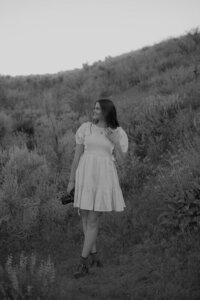 Black and white brand photo of Brittney standing on a hill. She's looking candidly to the side while holding her camera down at her side.