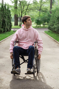 A young, masculine presenting person wearing a pink sweatshirt and jeans rolls navigates their wheelchair down a tree-lined, paved path. They look up and over their left shoulder, toward the sky.