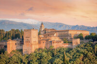 A scenic view of the Alhambra in Granada, Spain, with its historic architecture bathed in warm sunlight. The fortress is surrounded by lush greenery and set against a backdrop of distant mountains under a pastel-colored sky.