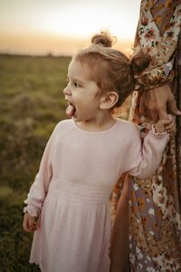 Young girl, dressed in a beautiful pink dress, adds a playful touch to the scene as she sticks out her tongue while holding her mom's hand. The mom is wearing a flower printed dress.
