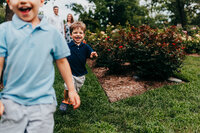 Young boy running and laughing towards the camera, with parents in the background.  Photo taken by Philadelphia family photographer, Kristi