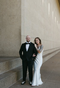 A couple holding onto one another next to a beige wall. 
