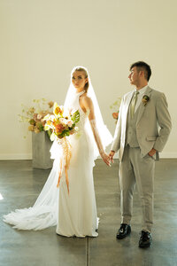 Wedding day photography in Rexburg Idaho and Idaho Falls. A bride holds the hand of her groom on their wedding day inside a simple venue. He is looking at her and she is looking at the camera with a neutral face.