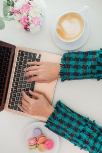 overview of white table with woman's hands on laptop with coffee, flowers and macaroons