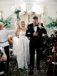 Kaitland and Kramer walk hand-in-hand down the aisle inside Boxwood Estate, surrounded by elegant indoor decor. The couple is smiling, with Kaitland wearing a flowing white gown and Kramer in a sharp suit. The venue features beautiful chandeliers and soft lighting, adding a romantic ambiance to the setting. Rows of white chairs are neatly arranged on either side of the aisle, and floral arrangements decorate the space, creating an intimate and sophisticated atmosphere.