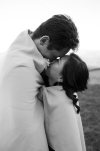 A bride and groom wrapped in a blanket stand in a lawn touching noses before a kiss captured by a Wilmington wedding photographer