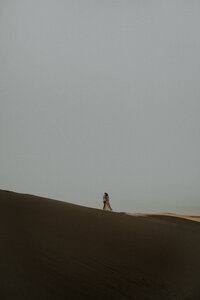 Couple stands, embracing each other, on top of a large sand dune at golden hour. It was shot from far away so it really emphasizes how large the sand dune and sky is all around them.