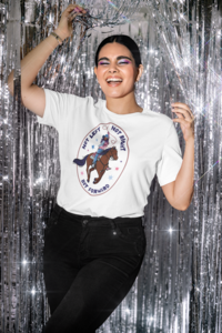A person wears a white T-shirt with an illustration of a cowgirl riding a horse and roping. The model is smiling and is standing in front of a shiny metallic silver party backdrop