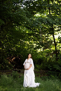 pregnant woman standing in a white gown, in a forest setting holding her pregnant belly looking down in awe.