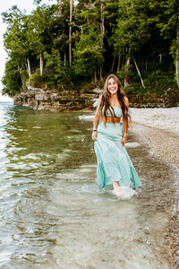 stunning senior girl with long brown hair in a two piece teal dress splashing her feet and laughing for her Green Bay senior photography session