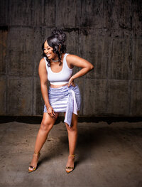 Discover BB's vibrant look featuring a purple sequin Miss Lola skirt paired with a stylish white tank top. Captured in a joyful moment against a cement wall, this pinup hairstyle by Pretty Faces by Chelsea adds the perfect touch of glam.