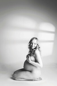 A gorgeous, modern, greyscale maternity photo captured by Mobile AL Maternity Photographer. The expecting mom poses nude by resting on her knees in the studio floor. The high key light perfectly highlights her feminine curves and growing belly. There is an artist window light overlay on the backdrop.