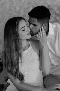 Engaged couple sits together on the sand at Jetty Park Beach while he kisses her cheek and she smiles, captured by an Orlando Proposal Photographer.