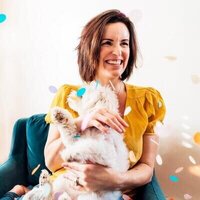 Gina Cooperman smiling and cuddling with a fluffy white dog among falling confetti.