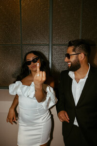 bride holding up her fourth finger to the camera while groom smiles at her