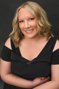 A professional portrait of a woman over 40 smiling woman with wavy blonde hair, wearing a black dress against a dark background. Captured with expert lighting and posing by Leslie Q Photography.