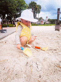 Baby with a life jacket on and sun hat playing in the sand.