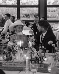 Black and white image of a smiling couple at a wedding reception. The woman wears a cowboy hat, surrounded by flowers and candles, creating a joyful atmosphere.
