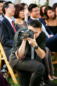 Photographer Henry Chen kneels to take a photo at a wedding dressed in black dress shirt and slacks