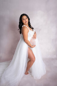 Woman looking over her shoulders, gently cradles her belly while accentuating her long tan leg through the slit of her maternity gown.
